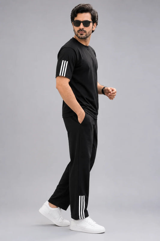 A man modeling a black two-piece tracksuit with white three-stripe accents on the sleeves and pants, paired with white sneakers, against a solid gray background.