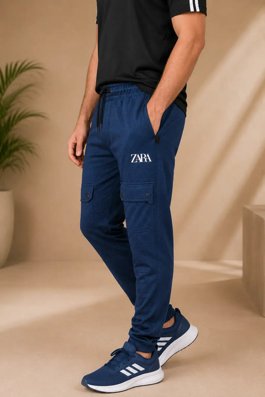 A man wearing navy blue cargo jogger pants with a white embroidered 'ZARA' logo on the left thigh, paired with a black t-shirt and navy Adidas sneakers, standing in a beige studio setting.