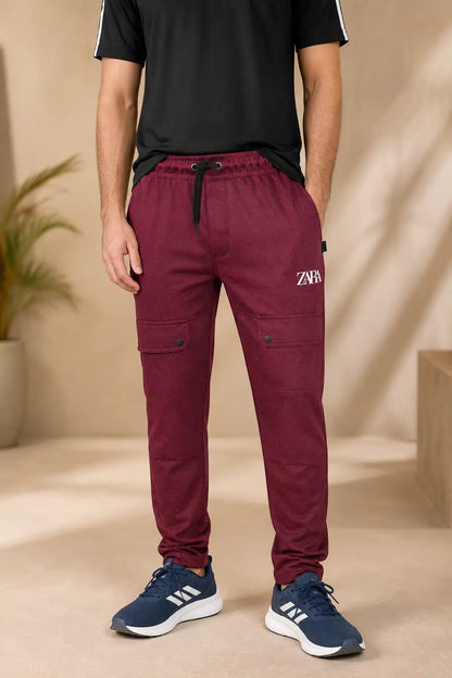 A person wearing burgundy jogger-style cargo pants with a drawstring waist and side pockets, paired with a black t-shirt and navy sneakers, standing in a warm beige indoor setting.