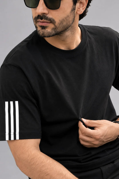 A man wearing a black short-sleeve t-shirt with three white stripes on the sleeve, photographed against a plain gray background.