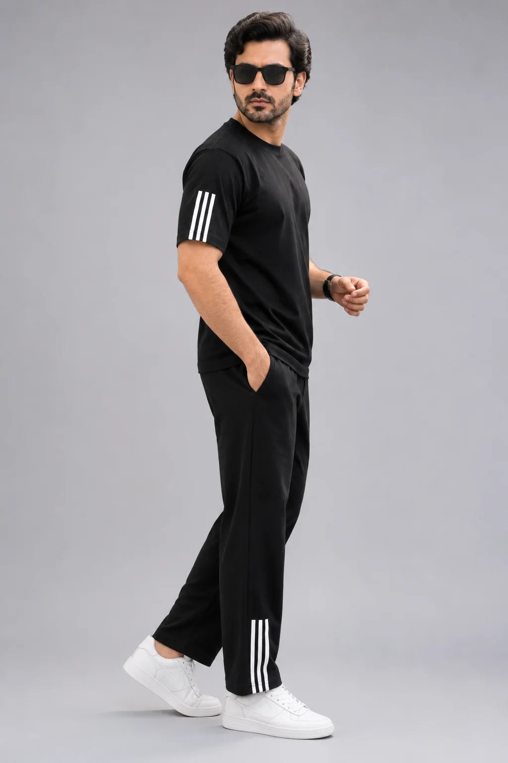 A man modeling a black two-piece tracksuit with white three-stripe accents on the sleeves and pants, paired with white sneakers, against a solid gray background.