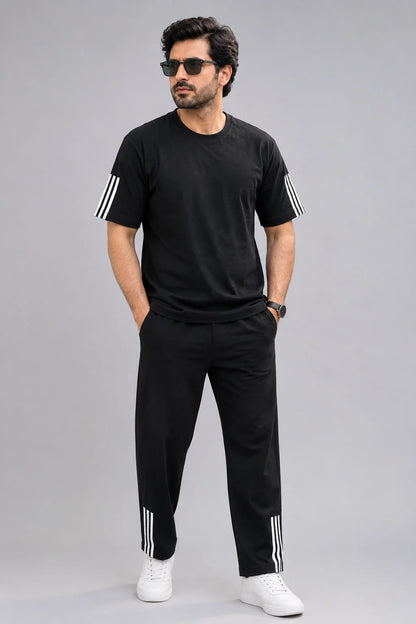 A man modeling a black short-sleeve t-shirt and matching black track pants, both featuring white stripes on the sleeves and lower legs, paired with white sneakers against a solid gray background.