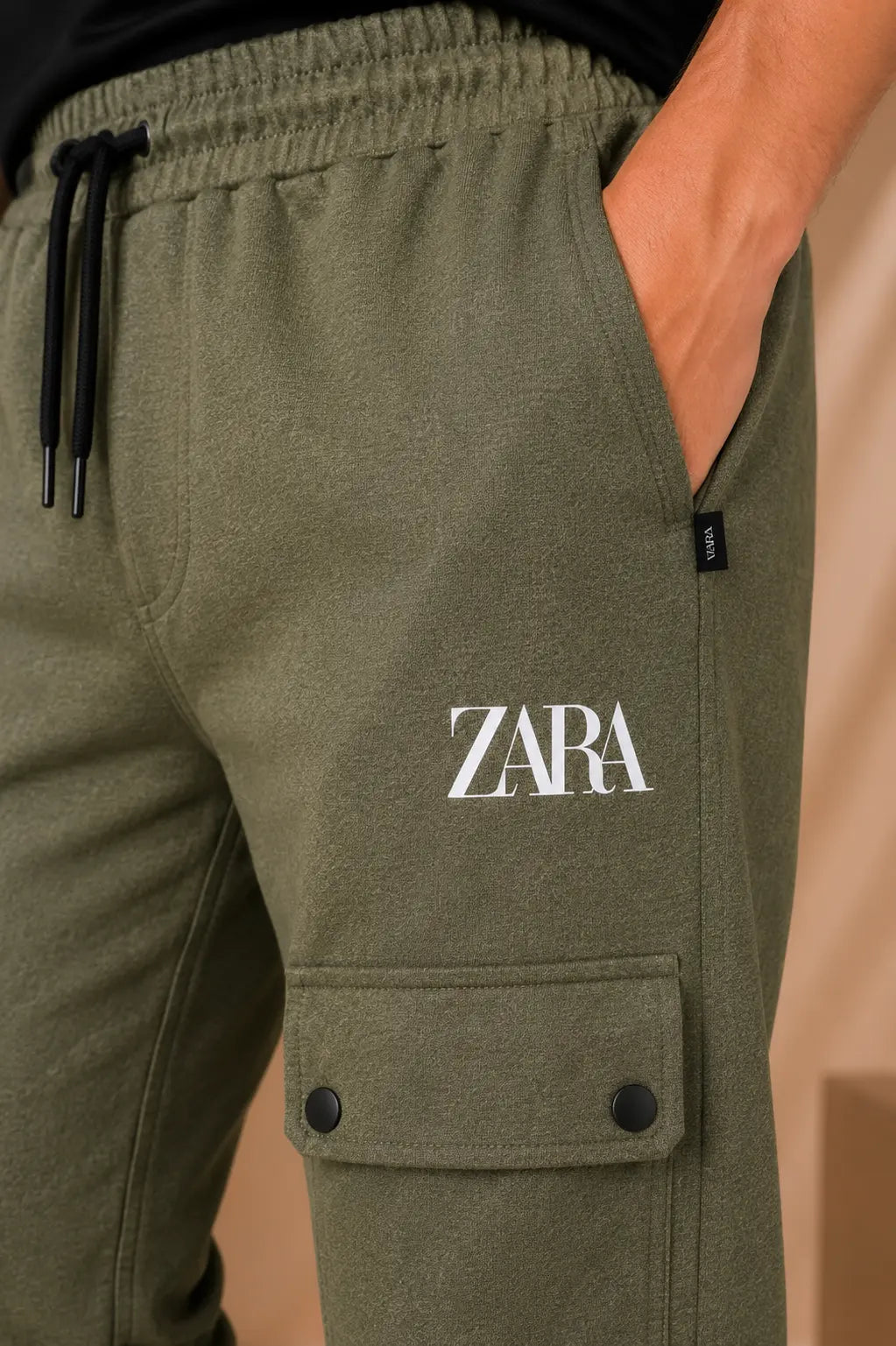 Close-up of olive green sweatpants worn by a model, featuring a white 'ZARA' logo printed on the thigh, a black drawstring waist, and a cargo pocket with snap buttons.