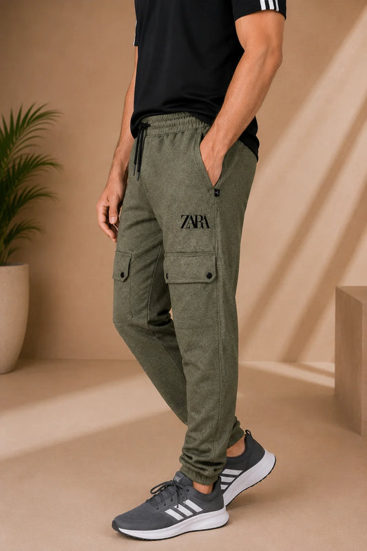 A man wearing olive green cargo jogger pants with a black drawstring waist, side cargo pockets, and a black embroidered logo on the thigh, paired with a black t-shirt and gray sneakers in a warm beige studio setting.