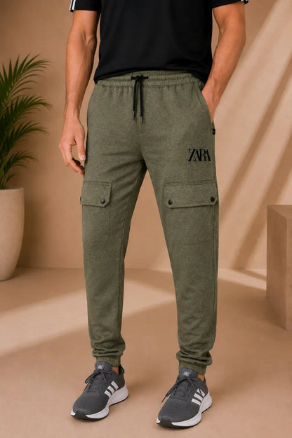 A man wearing olive green cargo jogger pants with a black drawstring and black logo on the thigh, paired with a black t-shirt and gray sneakers, in a warm beige studio setting.