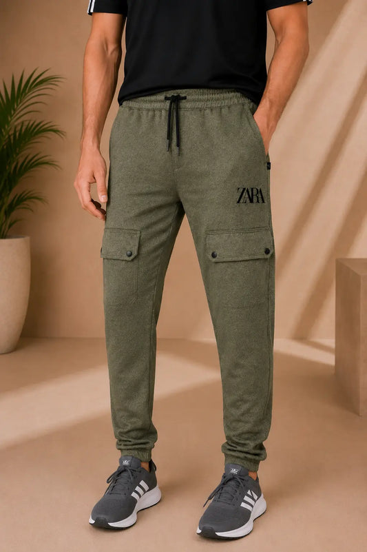 A man wearing olive green cargo jogger pants with a black drawstring and black logo on the thigh, paired with a black t-shirt and gray sneakers, in a warm beige studio setting.