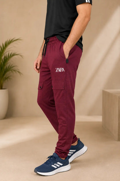 A person wearing burgundy jogger cargo pants with a white 'ZNA' logo on the left thigh, paired with a black t-shirt and navy blue sneakers, standing in a beige indoor setting.