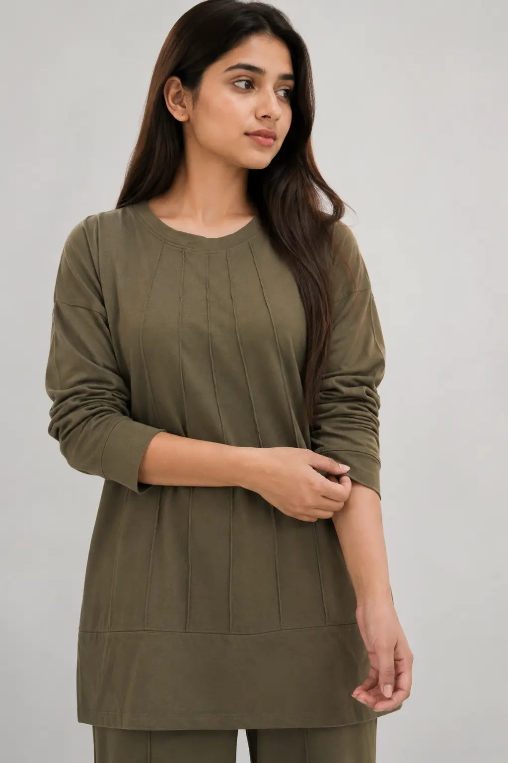 A woman wearing an olive green long-sleeve tunic top with vertical pleats, shown against a plain light gray studio background.