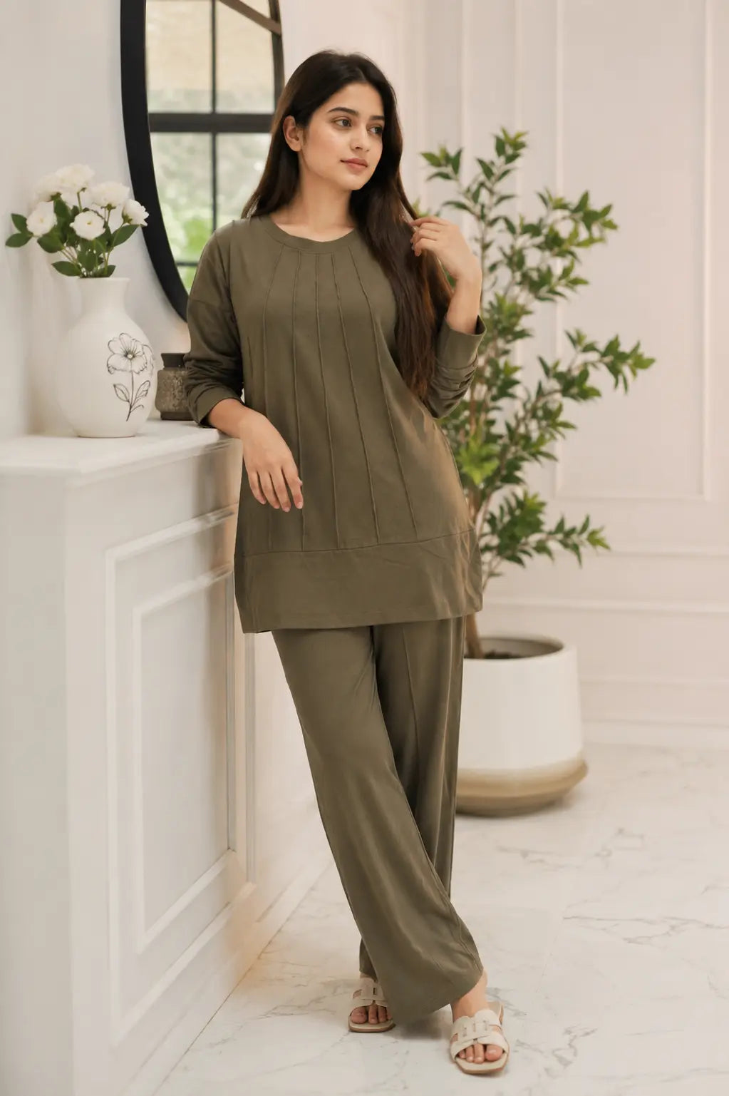 A woman modeling a matching two-piece lounge set in olive green, featuring a long-sleeve top with vertical pleats and wide-leg trousers, posed in a bright, modern interior setting.