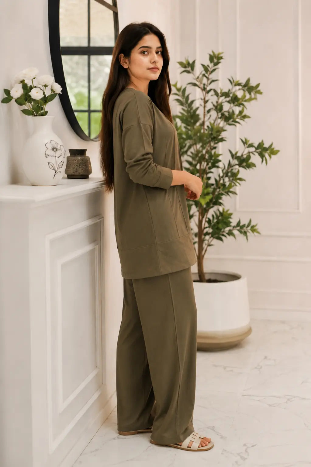 A woman modeling a matching two-piece lounge set in olive green, consisting of a long-sleeve top and wide-leg trousers, standing in a bright, modern room with white walls, a potted plant, and a white mantelpiece.