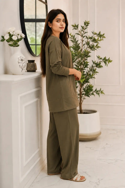 A woman modeling a matching two-piece lounge set in olive green, consisting of a long-sleeve top and wide-leg trousers, standing in a bright, modern room with white walls, a potted plant, and a white mantelpiece.