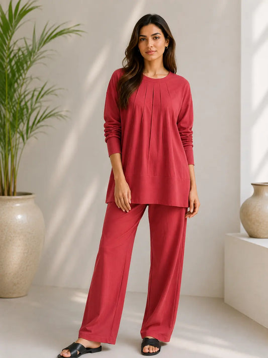 A woman modeling a matching two-piece lounge set in a warm red color, featuring a long-sleeve top with a gathered neckline and wide-leg pants, set against a minimalist beige interior with a potted plant.